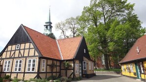 En traditionel bindingsværksbygning med rødt tegltag, et kirketårn i baggrunden og en belagt gårdsplads.
