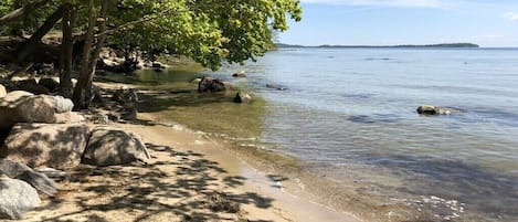 Ein Strand mit klarem Wasser, Felsen und einem Baum, der Schatten spendet.