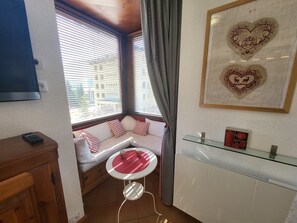 A cozy room with a window seat, a small table with a red mat, and a wall-mounted shelf with decorative items. - Central apartment, balcony, south/east view (Montgenèvre)