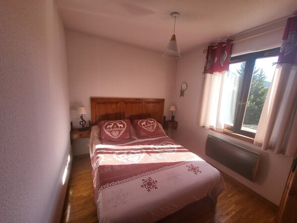A bedroom with a wooden headboard, a bed with heart-patterned pillows, a bedside table with a lamp, a window with curtains, and a radiator. - Central apartment, balcony, south/east view (Montgenèvre)