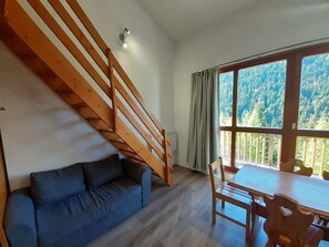 Een kamer met een houten trap, een blauwe bank, een eettafel met stoelen en een groot raam met uitzicht op een bos.