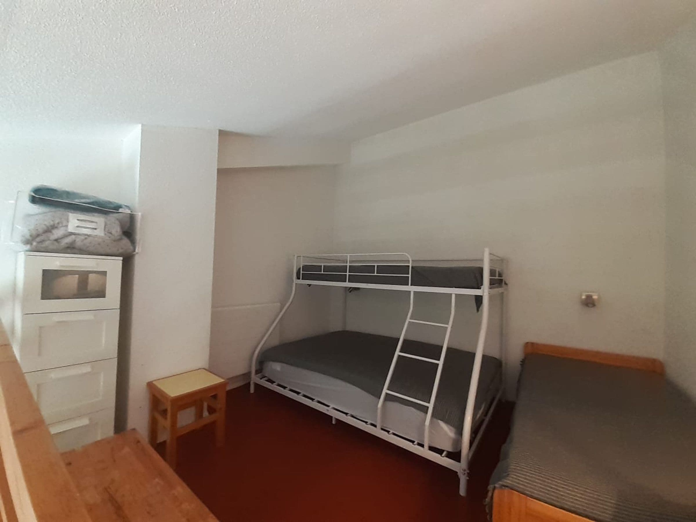 A small, single-bedded room with a bunk bed, a wooden desk, and a small stool.