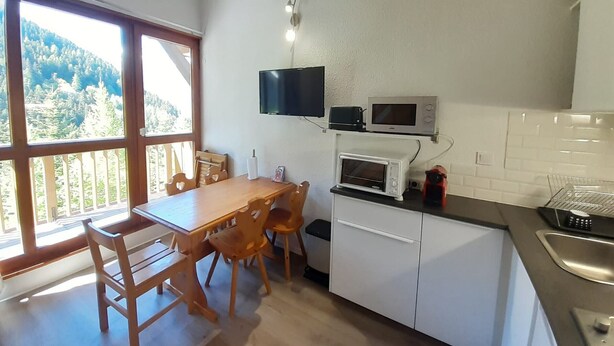 Eine Küche mit Esstisch, Stühlen, einem an der Wand montierten Fernseher und Blick ins Freie durch große Fenster.