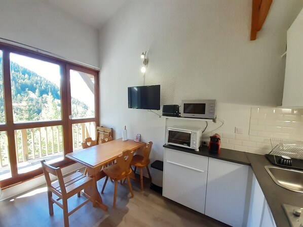Een keuken met een eettafel, stoelen, een televisie boven de toonbank en uitzicht op de omgeving door grote ramen.