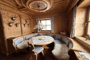 Interior de madeira com mesa redonda e assentos, teto decorativo e janela com cortinas.