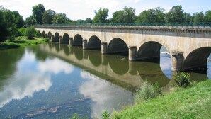 A stone bridge with multiple arches spanning a calm river, surrounded by greenery and a clear sky.