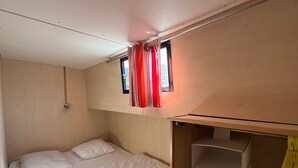 Een kleine kamer met een stapelbed, een klein raam met gordijnen, en een ingebouwde opbergkast met planken.