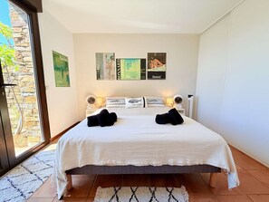 Habitación de hotel con cama, dos lámparas de noche, ventana con vista a un muro de piedra y una obra de arte en la pared.