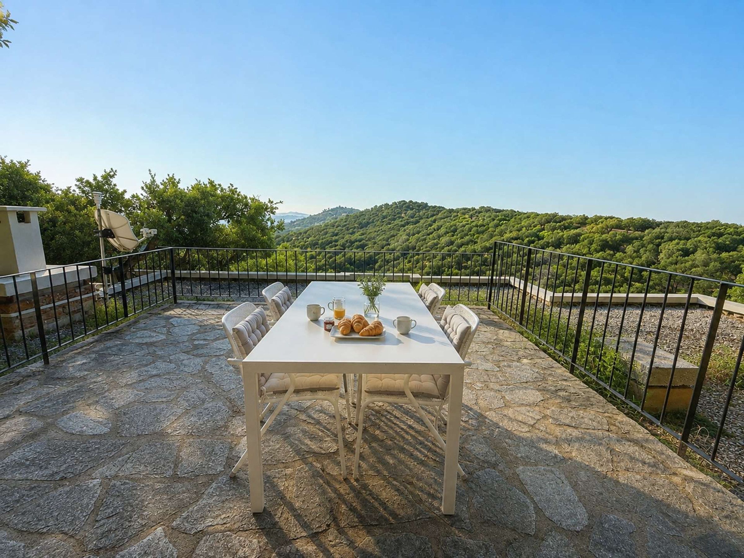 Une terrasse en pierre avec une table blanche dressée pour le petit-déjeuner, entourée de chaises, et offrant une vue sur un paysage verdoyant et luxuriant.