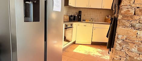 Ett kök med ett kylskåp i rostfritt stål, vita skåp, en vägg klädd med sten och ett klinkergolv.