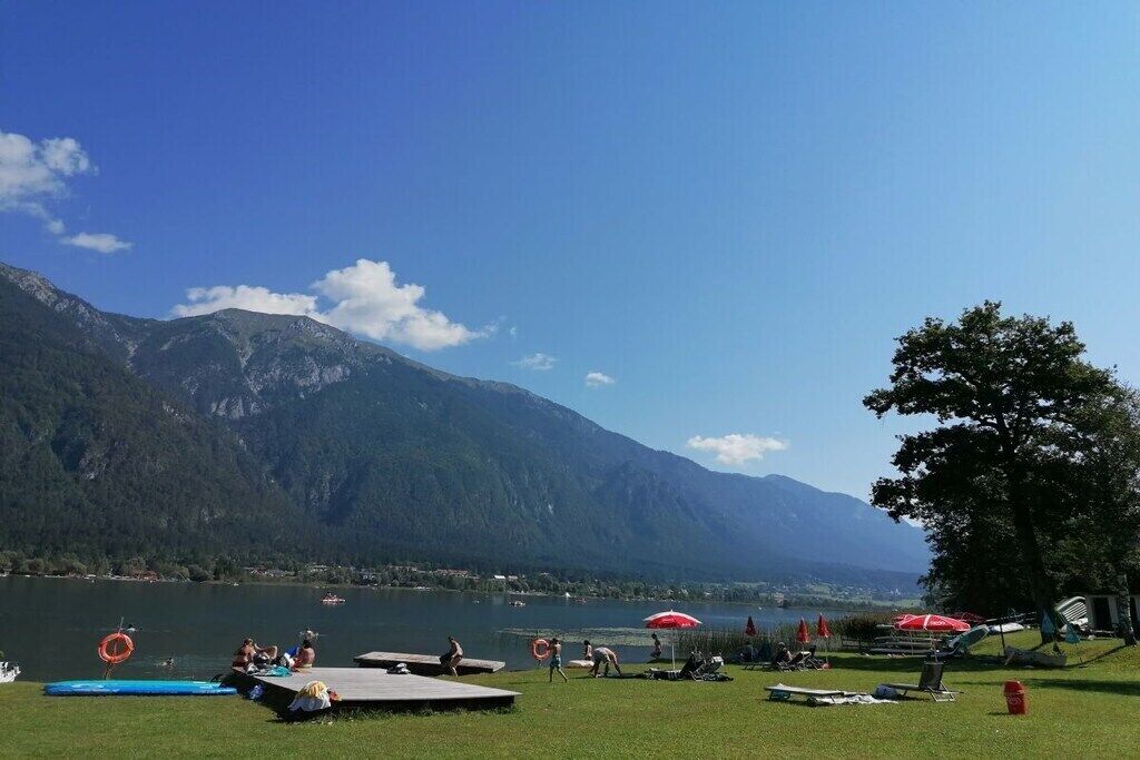 Vista del lago con montañas al fondo, un área verde con tumbonas y un cielo despejado.