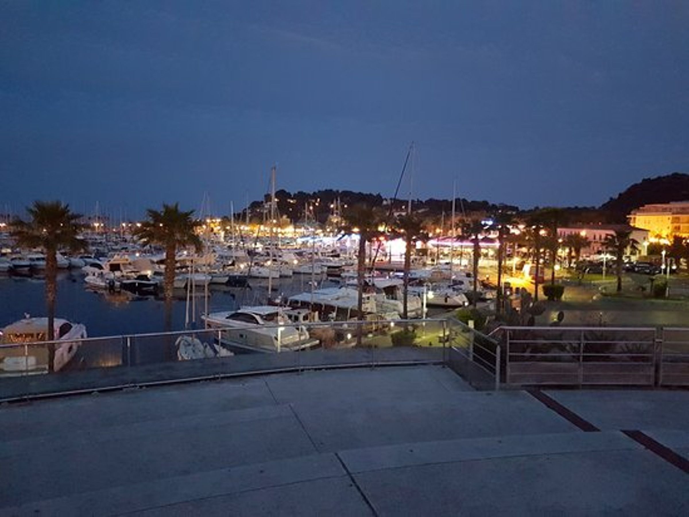 A marina at dusk with boats docked, a waterfront promenade, and buildings in the background.