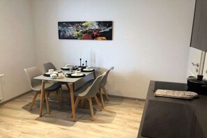 Een eetgedeelte met een tafel voor vier personen, stoelen, een schilderij aan de muur en een zwarte bank met een kussen.