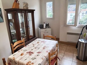 A dining area with a table covered in a floral tablecloth, wooden chairs, a cabinet with glass doors, a window with a view of buildings, and a radiator. - Apartment with parking in city center - 3 guests (Amélie-les-Bains-Palalda)