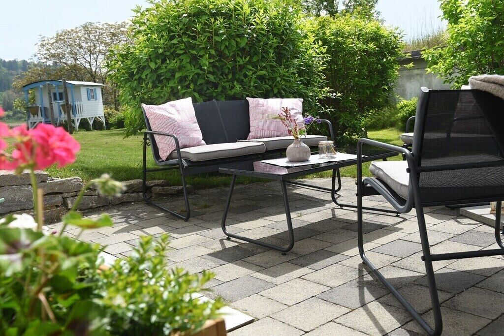 An outdoor patio with a sofa and chairs, a small table with a vase, and a blue house in the background.