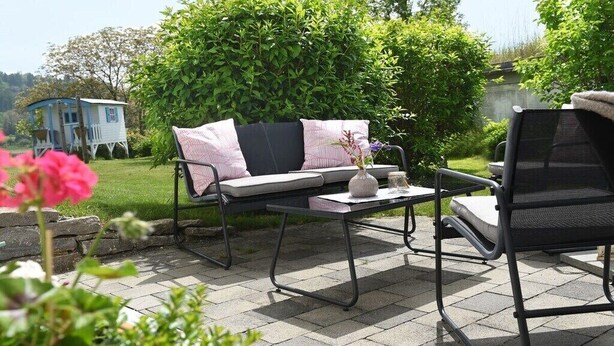 En udendørs terrasse med en sofa og stole, et lille bord med en vase, og et blåt hus i baggrunden.