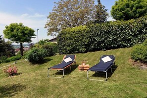 Zwei Liegestühle mit Kissen auf einer grünen Wiese, umgeben von Pflanzen und einer Hecke.
