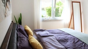 Et soveværelse med seng, et vindue med gardiner, et gulvspejl og et billede på væggen.