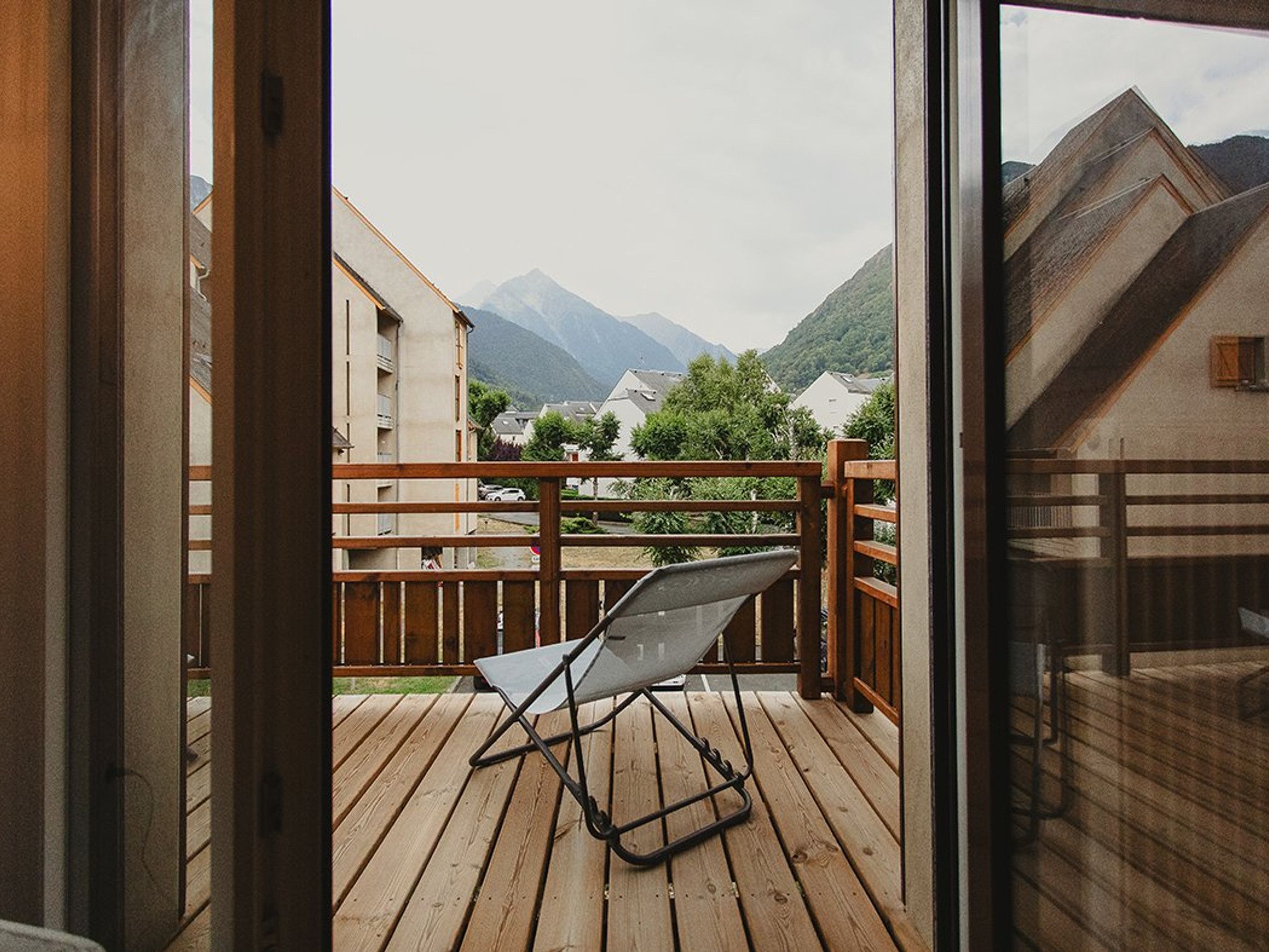A balcony with a metal chair, wooden railing, and a view of buildings and mountains.