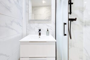 A modern bathroom with a white marble vanity, a glass shower enclosure, and a large mirror. - Parisian Studio Near Les Halles & Marais (Paris)