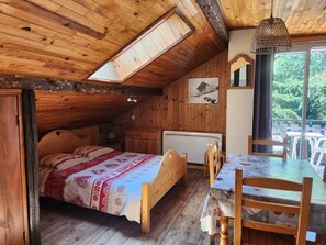 Uma cabana acolhedora com teto inclinado, uma cama com colcha estampada, uma mesa de jantar com cadeiras e vista para o verde lá fora.
