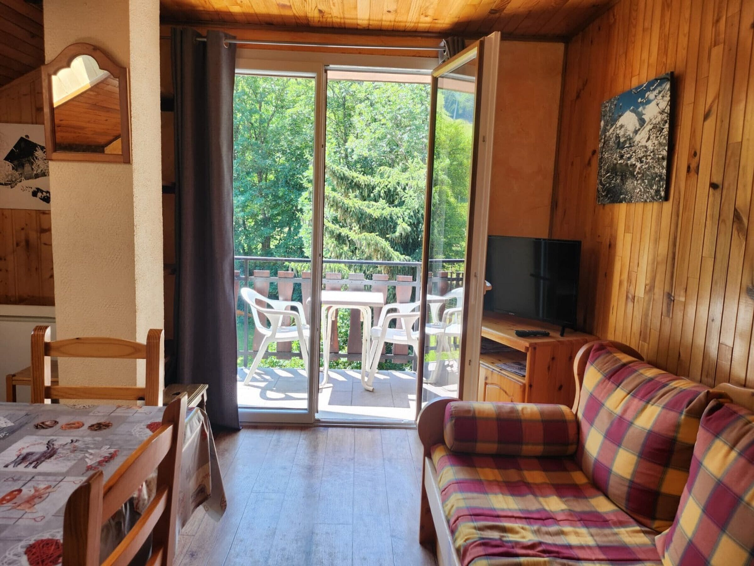 A room with a wooden interior, a sofa, a table with chairs, a television, and a sliding glass door leading to an outdoor area with a table and chairs.