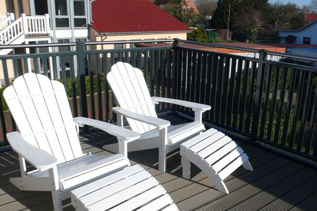 Dos sillas Adirondack blancas sobre una terraza de madera con barandilla negra.