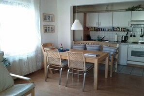 A dining area with a wooden table and chairs, a kitchen in the background, and a window with curtains. - Holiday apartment in Rerik with swimming pool (Rerik)