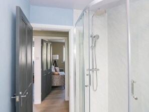 A modern bathroom with a glass shower enclosure, a wooden door, and a hallway leading to a bedroom with a bed and a lamp. - Aqueduct Residence (Lancaster)