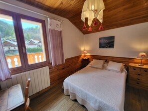 A bedroom with a wooden ceiling, a bed with white bedding, a nightstand with a lamp, a window with curtains, and a radiator. - Cozy apartment with parking - 10 guests (Valloire)