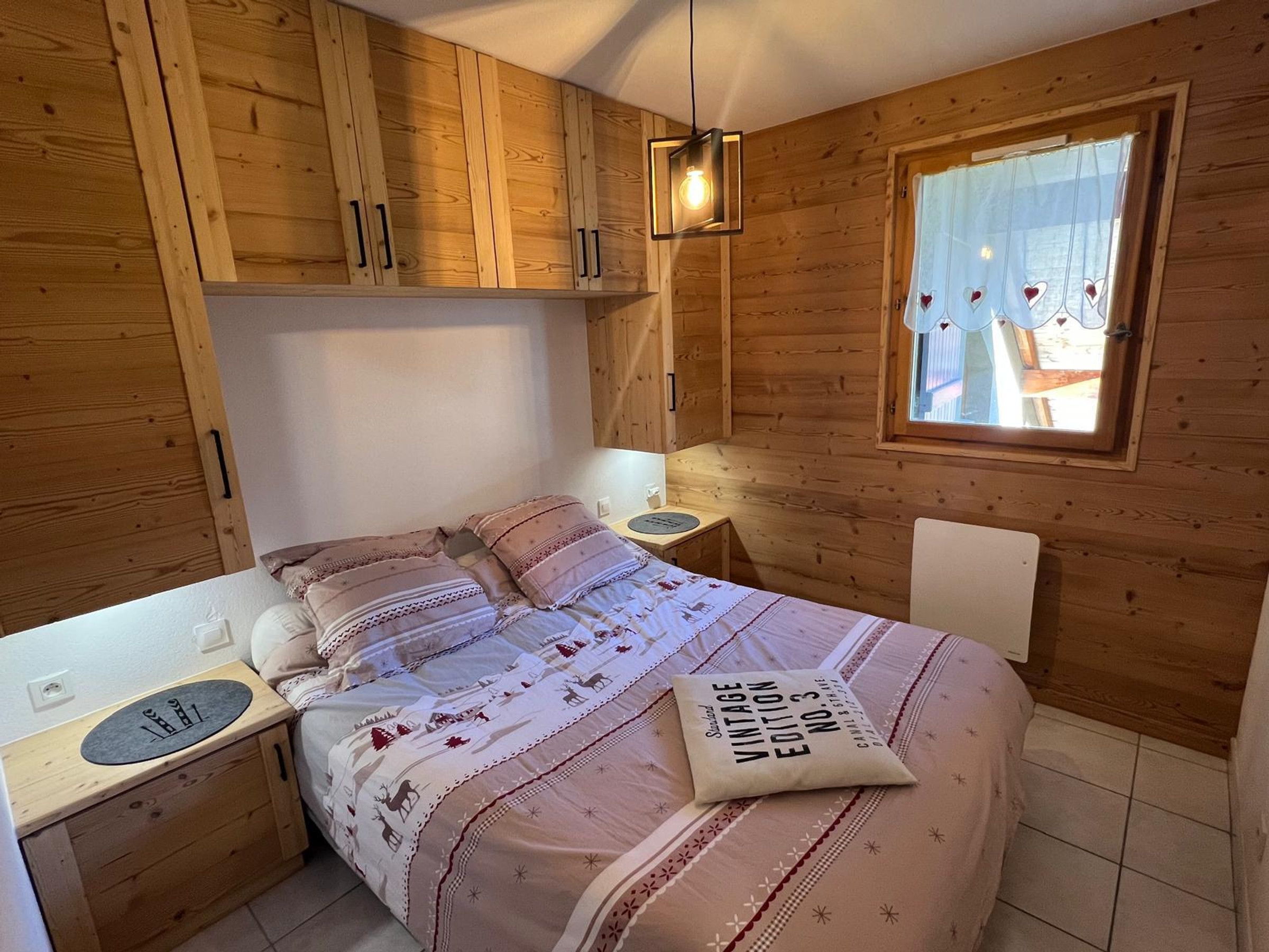 Un dormitorio acogedor con paredes de madera, una cama con ropa de cama estampada, una mesita de noche con una lámpara y una ventana con cortinas.