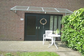 A brick building with a black door, a numbered plaque, a white chair, a small table, a potted plant, and a green hedge. - Aquadelta De Tong 155 (Bruinisse)