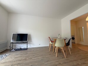 A room with a TV on a stand, a dining table with chairs, and a washing machine in the background. - Charming apartment by the beach - 3 guests (Arcachon)