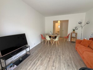 A living room with a flat-screen TV, a dining area with a table and chairs, and an orange sofa. - Charming apartment by the beach - 3 guests (Arcachon)