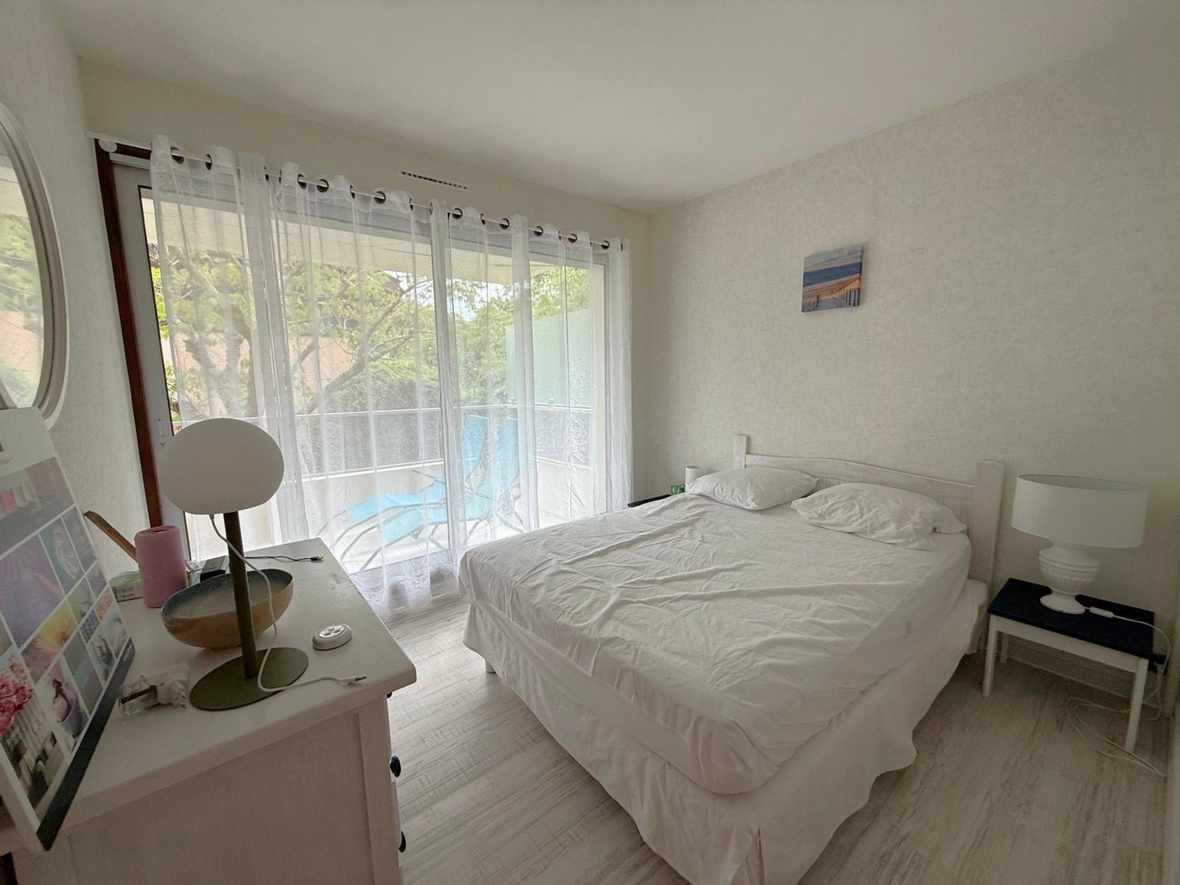 A bedroom with a bed, a bedside table with a lamp, a desk with a mirror, and a window with curtains.
