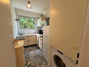A compact kitchen with a washing machine, a window with a view of greenery, and a patterned floor. - Charming apartment by the beach - 3 guests (Arcachon)