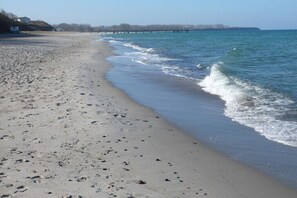 Una playa con un muelle que se adentra en el agua, orilla arenosa y cielo despejado.