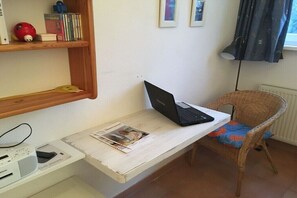 Ein Zimmer mit einem Laptop auf einem weißen Schreibtisch, einem Korbstuhl, einem Regal mit Büchern und einem roten Spielzeug sowie einem Heizkörper.