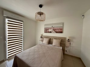 A bedroom with a bed, a hanging light fixture, a wall-mounted picture, and a window with blinds. - Modern house near forest - PMR - 6 guests (Saint-Jean-de-Monts)