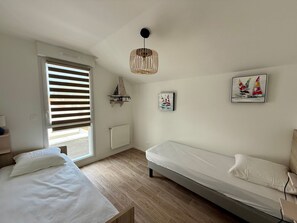 Ein Schlafzimmer mit einem Bett, einem Fenster mit Jalousien, einem Heizkörper, einer Pendelleuchte und zwei Wandbildern.