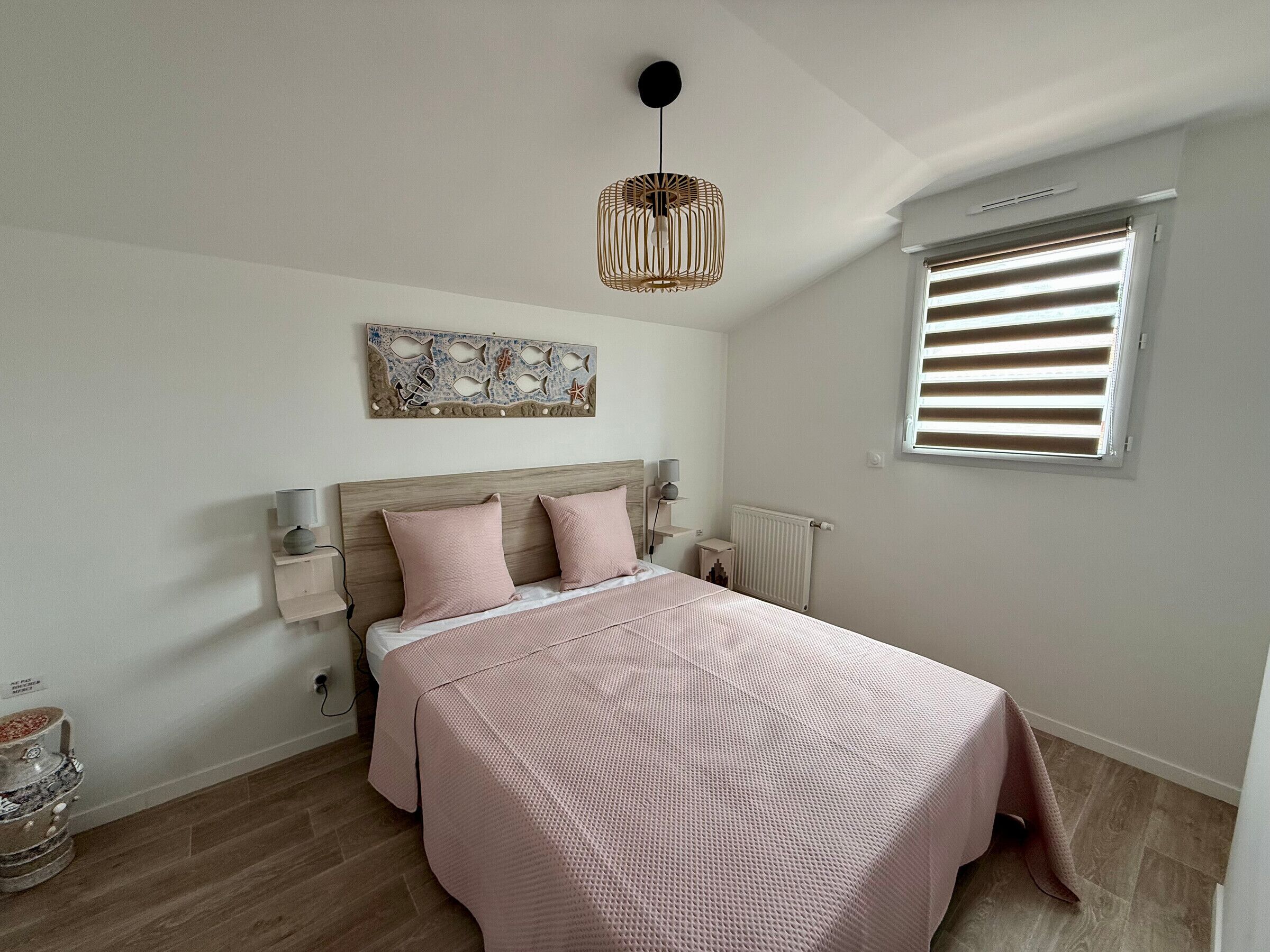 A bedroom with a bed, a bedside table, a wall-mounted light fixture, a window with blinds, and a decorative wall piece.