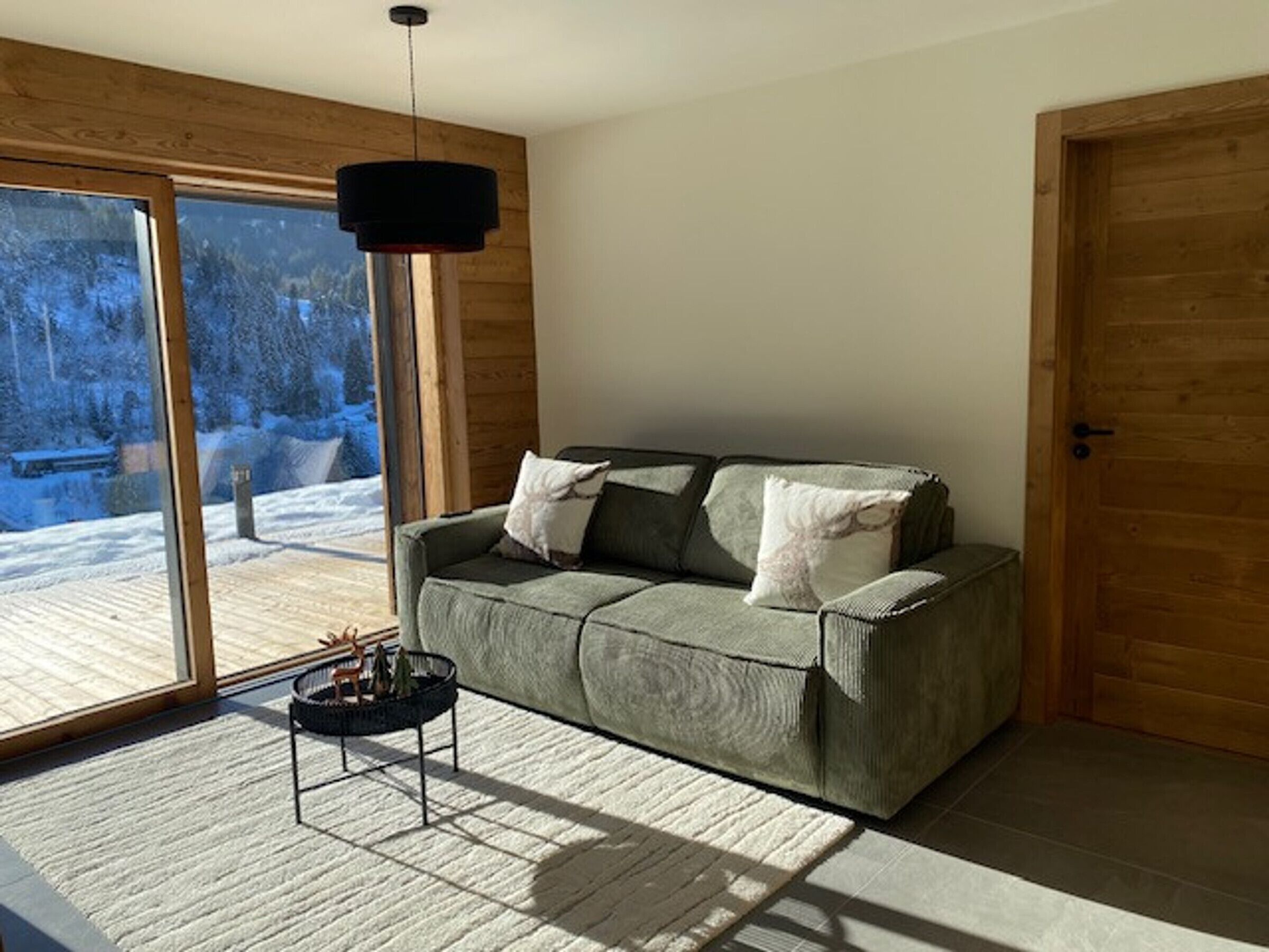 Una sala moderna con un sofá verde, una lámpara colgante negra y vistas a un paisaje nevado a través de amplios ventanales.