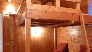 Un intérieur en bois avec une structure de lit superposé, un petit tabouret et un luminaire fixé au mur.