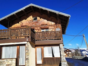 A wooden chalet with a balcony, stone walls, and a mountain backdrop.
