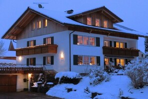 Un chalet de dos pisos con techo nevado, varios ventanales y una escalera de piedra que conduce a la entrada.