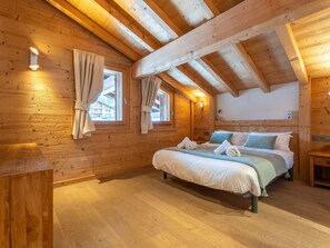 Ein Schlafzimmer einer Holzhütte mit einem Bett, zwei mit Vorhängen versehenen Fenstern, einer Holzbalkendecke und einem kleinen Nachttisch.