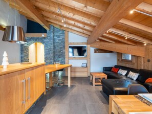 A modern kitchen with a stone wall, wooden ceiling beams, and a black leather sofa in a living area. - Ideal family apartment - 6 guests (Les Gets)