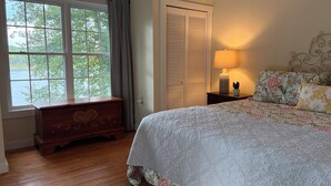 Un dormitorio con un aparador de madera, una cama con ropa de cama floral, una mesita de noche con una lámpara y una ventana con vistas a árboles.