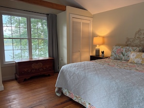 A bedroom with a wooden dresser, a bed with floral bedding, a nightstand with a lamp, and a window with a view of trees.