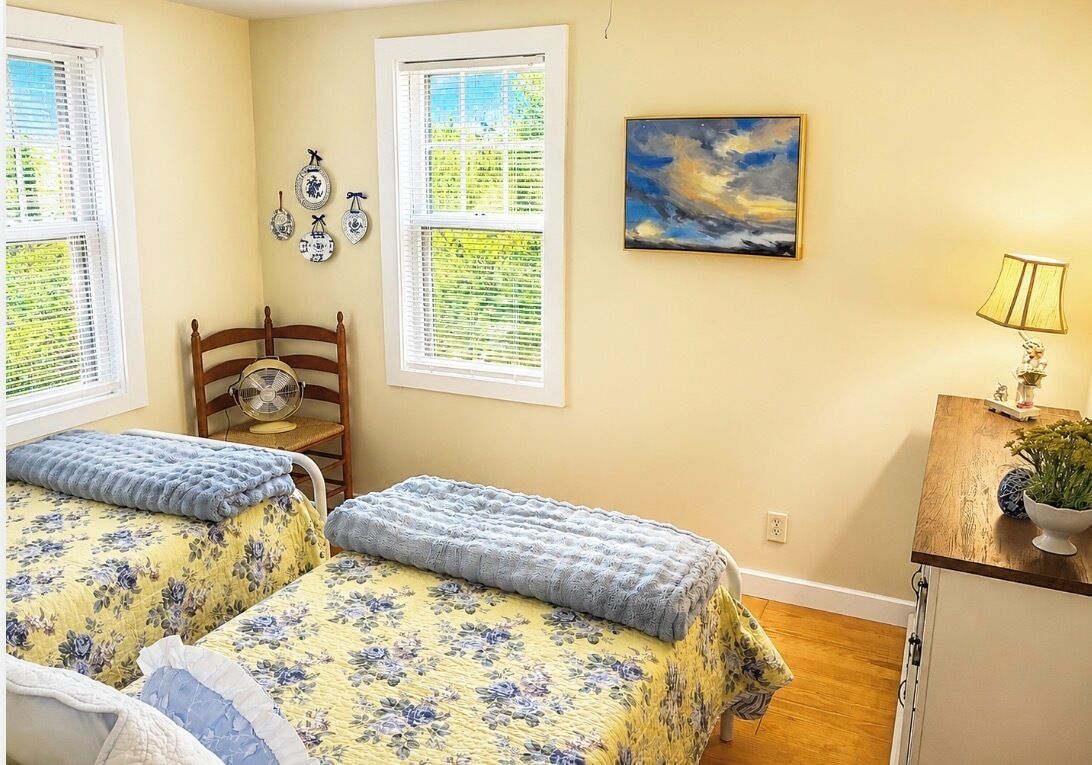 A room with two beds, a wooden chair, a table with a lamp, and a painting on the wall.